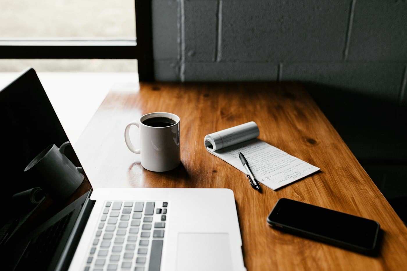 A laptop, a mug of coffee and a handwritten notepad on a wooden desk by a window
